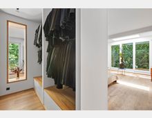 Ein modernes Schlafzimmer mit angrenzendem Ankleidebereich. Der Raum hat helle Holzböden, eine große Fensterfront mit Blick auf eine begrünte Wand und ein schlichtes, weißes Interieur.