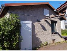 Ein kleines Gebäude mit grober, beigefarbener Putzfassade, einem roten Satteldach und einer weißen Tür. Neben der Tür ist ein Wandlicht angebracht, und ein Fenster mit dunklem Rahmen befindet sich rechts daneben; davor wächst ein dichter Strauch.