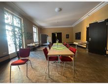 Ein einfacher Seminarraum mit Holzfußboden, mehreren aufgestellten Tischen, roten Stühlen und großen Fenstern auf der linken Seite. Pflanzen, Wandkunst und eine Tafel im Hintergrund ergänzen das helle, minimalistische Ambiente.