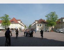 Eine gepflasterte Fußgängerzone mit historischen Gebäuden auf beiden Seiten, teils mit rot gedeckten Dächern. Menschen flanieren, sitzen in Straßencafés und fahren Fahrrad, umgeben von grünen Bäumen und hellem Himmel.