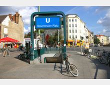 U-Bahnhofseingang Rosenthaler Platz in Berlin, umgeben von einer städtischen Szene mit historischen und modernen Gebäuden, einem Café mit roten Sonnenschirmen und Fahrrädern, die an einem Geländer abgestellt sind. Eine belebte Straßenkreuzung ist im Hintergrund zu sehen.