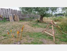 Ein Bogenschießplatz im Freien mit grüner Wiese, Holzständer für Bögen und Pfeile in einem Block. Im Hintergrund steht ein Holzzaun, mehrere Zielscheiben und ein Baum, umgeben von natürlicher Vegetation.
