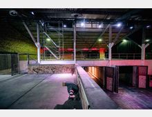 Industriehalle mit unverputzten Ziegelwänden, Metallträgern und Betonelementen. Im Vordergrund eine erhöhte Plattform mit einer kleinen Rampe, im Hintergrund Gerüste, offene Tore und rote Lichtinstallationen.