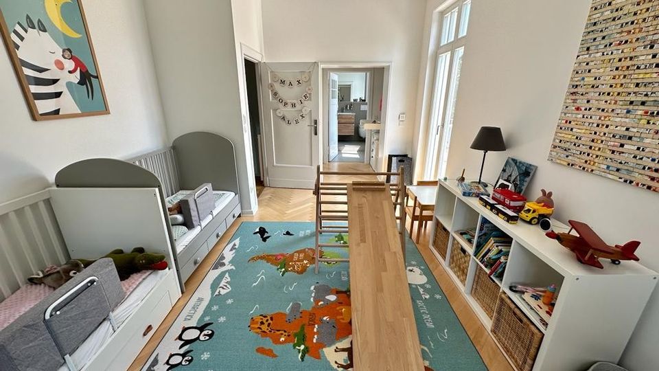 Helles Kinderzimmer mit zwei Betten an der linken Seite, einem bunten Teppich mit Tiermotiven auf dem Boden und einer langen Holzrampe in der Mitte. Rechts befinden sich Regale mit Spielsachen und Büchern, sowie ein kleiner Spieltisch vor einer großen Glastür.