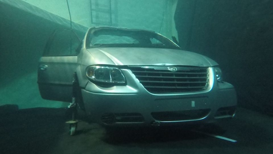 Underwater scene showing a silver minivan inside a submerged tunnel or parking structure, with concrete walls and a ladder visible in the background. The vehicle is partially tilted on the floor, surrounded by murky water and scattered debris.