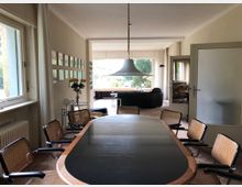 Modernes Interieur eines Wohnbereichs mit einem Besprechungstisch aus Holz und schwarzer Oberfläche, umgeben von Stühlen mit Korbgeflecht. Im Hintergrund ein Wohnzimmer mit großen Fenstern, die auf einen Garten blicken, sowie eine Hängelampe und weitere Sitzgelegenheiten.