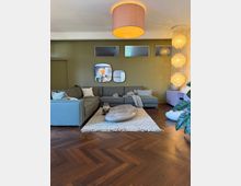 Ein modern eingerichtetes Wohnzimmer mit einer großen, grauen Ecksofa-Gruppe und einem flachen, ovalen Couchtisch auf einem cremefarbenen Teppich. Der Raum hat eine olivgrüne Wand mit kleinen Fenstern und Spiegeln, einen hölzernen Fischgrätboden, eine hängende dekorative Lampeninstallation und eine große Topfpflanze am Rand.