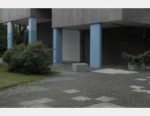 Außenbereich eines modernen Gebäudes mit grauer Fassade, blauen Stützpfeilern und einem gepflasterten Vorplatz. Links ein grüner Busch und rechts kleinere Betonpflanzkästen mit Sträuchern.