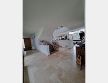 Offener, moderner Wohnbereich mit hellen Fliesenböden, einer freistehenden geschwungenen Treppe mit schwarzem Metallgeländer, großen Fenstern mit Blick ins Grüne und dekorativen Elementen wie einer Akustikgitarre und blühenden Pflanzen.