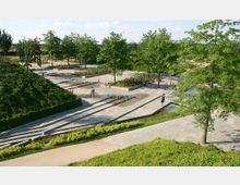 Ein moderner Park mit angelegten Grünflächen, Bäumen und schattigen Spazierwegen. Im Zentrum befindet sich ein flaches Wasserspiel mit kleinen Fontänen, umgeben von terrassierten Steinstufen und bepflanzten Beeten.