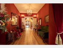 Ein eleganter Flur mit roten Wänden und Holzboden, dekoriert mit antiken Möbeln, Blumenarrangements und einem Kristallleuchter. Links und rechts stehen gepolsterte Stühle und Tische, während im Hintergrund eine geöffnete Doppeltür den Blick in einen weiteren Raum mit einem großen Fenster freigibt.