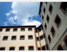 Blick auf die Fassaden eines historischen Gebäudekomplexes mit mehreren Stockwerken, gekennzeichnet durch helle Putzwände, rote Ziegelrahmen um die Fenster und vergitterte Fenster. Der Himmel ist teils bewölkt und bildet den Hintergrund.