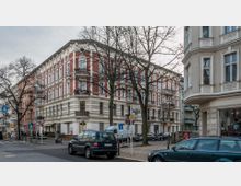 Straßenecke mit mehrstöckigen Altbauwohnungen im Gründerzeitstil, gekennzeichnet durch rote und beige Fassaden mit weißen Stuckverzierungen. Im Vordergrund geparkte Autos, Verkehrszeichen, Fußgängerbereich und Bäume ohne Laub.