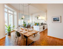 Heller, offener Wohnraum mit Essbereich, ausgestattet mit einem langen Holztisch, modernen Stühlen und hängenden Glühbirnenlampen, daneben eine helle Einbauküche mit Pflanzen und Dekor auf der Arbeitsfläche. Große Fenster lassen viel Tageslicht herein, und Pflanzen sowie Holzböden sorgen für ein warmes Ambiente.