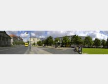 Weitläufiger Platz in einer Stadt mit gepflasterten Wegen, einem zentralen Brunnen und Grünflächen, umgeben von Bäumen und mehrstöckigen Gebäuden im Hintergrund. Links fährt ein gelber Bus die Straße entlang, während Passanten den Platz beleben.