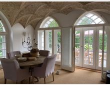 Wohnraum mit historischem Gewölbedecke und floralen Verzierungen, großen weißen Sprossenfenstern und Glastüren, die zu einer Terrasse mit Holzmöbeln führen. In der Mitte steht ein runder Holztisch mit hellen Polsterstühlen und einem dekorativen Kerzenständer.