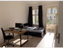 Helles Schlafzimmer mit Holzboden, ausgestattet mit einem Schreibtisch, Bürostuhl, Computer und einem einzelnen Bett. Große Fenster mit dunklen Vorhängen bieten Blick auf einen Balkon mit Stuhl und Tisch sowie auf ein gegenüberliegendes Gebäude.