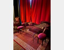 Ein eleganter Innenraum mit einem roten Samtsofa und zwei gepolsterten Hockern vor einem Glastisch, der auf einem roten Teppich mit Muster steht. Im Hintergrund hängen schwere rote Vorhänge, links steht ein Holzstuhl mit Polster.