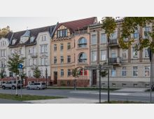 Straßenansicht von Wohnhäusern im architektonischen Stil der Gründerzeit mit mehreren Etagen, verzierten Fassaden und Erkern. Im Vordergrund befinden sich eine Straße mit parkenden Autos, Bäume sowie ein Straßenschild mit den Beschriftungen „Buschmühlenweg“ und „Carthausplatz“.