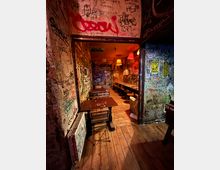 Schmaler Bar- oder Kneipeninnenraum mit Holzfußboden, kleinen Tischen und Bänken sowie einer langen Sitzbank mit Hockern entlang der rechten Wand. Wände, Decke und Türen sind dicht mit Graffiti und Aufklebern bedeckt, beleuchtet von warmen, hängenden Lampenschirmen.
