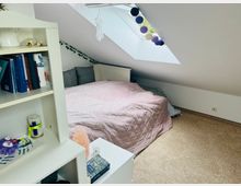 Ein Dachzimmer mit einem Bett, das unter einer schrägen Decke mit Dachfenster steht. Neben dem Bett steht ein weißes Regal mit Büchern und Dekorationen, der Boden ist mit Teppich ausgelegt.