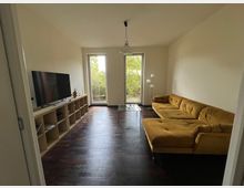 Wohnzimmer mit dunklem Holzboden, einer gelben Eckcouch, einem modernen TV auf einem offenen Regal und zwei bodentiefen Fenstern, die auf eine grüne Außenfläche blicken. Eine minimalistische Deckenlampe hängt in der Mitte des Raumes.