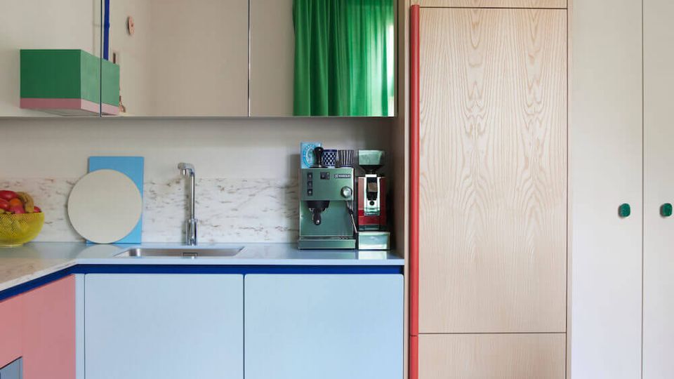 Modern compact kitchen with pale blue and pink lower cabinets, mirrored upper cupboards, and a marble backsplash with an integrated sink and countertop appliances. A tall light-wood pantry stands beside a white storage wall, with green curtains and a marble dining table with red chairs in the foreground.