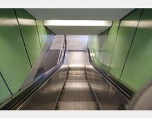 Rolltreppe in einem überdachten, engen Bereich mit grünen Wandverkleidungen auf beiden Seiten. Am unteren Ende der Rolltreppe führt der Weg zu einem Bereich mit hellem Bodenbelag.