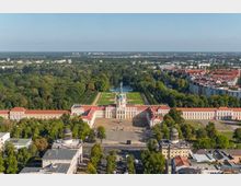 Luftaufnahme des Schlosses Charlottenburg in Berlin, einem barocken Gebäude mit rotem Ziegeldach und zentraler Kuppel, umgeben von symmetrischen Gartenanlagen und weitläufigem Park. Im Vordergrund sind Straßenzüge und Gebäude sichtbar, im Hintergrund erstreckt sich die Stadtlandschaft.