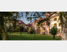 Wohngebäude mit hellgelber und roter Fassade im Stil der Gründerzeit, umgeben von einer grünen Parkanlage mit Rasenflächen und einzelnen, teils blattlosen Bäumen. Im Vordergrund werden einige Äste von Bäumen sichtbar, die den Blick rahmen.