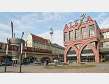 Platz mit Blick auf das Einkaufszentrum "Alexa" mit auffälliger Fassade in rosa Stein und großen Rundbögen, im Hintergrund der Berliner Fernsehturm. Rechts daneben befinden sich eine Hochbahntrasse mit S-Bahn und Gebäude im klassischen Baustil mit roten Dächern.