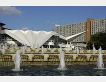 Modernes Gebäude mit markanter weißer, gefalteter Dachstruktur und großen Glasflächen im Vordergrund, umgeben von einem weitläufigen Brunnen mit mehreren Wasserspielen; dahinter ein mehrstöckiges Wohn- oder Bürogebäude mit rechteckigen Fenstern.