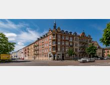 Eckgebäude in einer städtischen Straße mit historischer Fassade aus roten und beigefarbenen Elementen, vielen Fenstern und kleinen Balkonen. Im Vordergrund parkende Autos, Fahrräder und einige Bäume; im Hintergrund blauer Himmel mit wenigen Wolken.