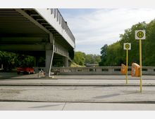 Bushaltestelle unter einer modernen Brücke mit breiten Stahlträgern, daneben ein parkendes rotes Auto und ein Fahrrad. Im Hintergrund sind Bäume und ein ruhiger Fluss zu sehen, während gelbe Haltestellenschilder und Mülleimer den Vordergrund prägen.