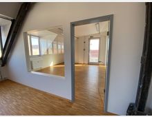 Moderner Innenraum eines Gebäudes mit Holzboden, schräger Decke und großen Fenstern, die viel Tageslicht einlassen. Sichtbar sind eine Glastür, ein quadratisches Innenfenster und ein Heizkörper unter den Fenstern.