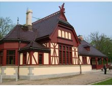 Historisches Gebäude im Fachwerkstil mit cremefarbenen Wänden und dunkelroten Holzelementen sowie einem Dach aus dunkelbraunen Ziegeln, umgeben von Bäumen und einem unbefestigten Untergrund.