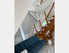 Innenraum eines modernen Wohnbereichs mit einem blauen Sofa im Vordergrund und einer Holztreppe im Hintergrund, die zu einer offenen Galerie führt. Die weißen Wände und Holzdetails verleihen dem Raum eine helle und minimalistische Atmosphäre.