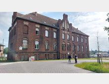Backsteinbau mit drei Stockwerken und Satteldach, typisch für historische Industriegebäude, umgeben von gepflasterten Wegen und spärlichem Grün. Im Vordergrund stehen zwei Menschen, während im Hintergrund Bahngleise und ein Schild sichtbar sind.