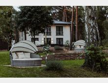 Moderne Gartenlandschaft mit einer weißen Villa im Hintergrund, umgeben von hohen Bäumen. Im Vordergrund befinden sich gepolsterte, runde Gartenliegen mit Sonnenschutz, umgeben von gepflegtem Rasen und Zierpflanzen.