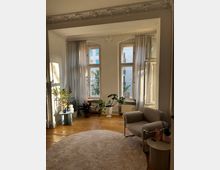 Ein helles Wohnzimmer mit hohen Decken und verzierten Stuckdetails. Drei große Fenster mit dünnen Vorhängen lassen Tageslicht herein, während Zimmerpflanzen, ein beigefarbener Teppich, eine Sessel und ein Beistelltisch den Raum schmücken.