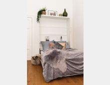 Kompaktes Schlafzimmer mit einem Holzfußboden, einem ausklappbaren Bett mit grauer Überdecke und Fellakzent, dekorativen Kissen und einem hohen weißen Regal darüber. Auf dem Regal befinden sich Pflanzen, gerahmte Bilder und Glasvasen.