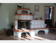 Gemütlicher Wohnbereich mit einem weißen, gekachelten Kamin mit Holzablage und Glasfront. Daneben stehen Körbe mit Holz und Zeitschriften; im Hintergrund sind Teil eines Sessels sowie eine Türöffnung sichtbar.