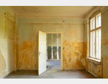 Unrenovierter Innenraum eines alten Gebäudes mit abblätterndem Putz und freigelegten Wandflächen, Holzboden und offener weißer Tür zu einem weiteren kahlen Raum. Rechts sorgt ein großes Sprossenfenster mit tiefer Laibung für Tageslicht.