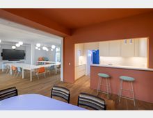 Ein moderner, offener Innenraum mit einem Besprechungsraum auf der linken Seite, ausgestattet mit langen weißen Tischen, bunten Stühlen und einer großen Wandbildschirm. Rechts befindet sich eine kleine Küchenzeile in einer Wand mit orangefarbener Akzentfarbe, einer hellen Arbeitsfläche, Hockern und schlichten weißen Schränken.