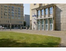 Großer Platz mit einer Grünfläche im Vordergrund und umliegenden Gebäuden im Hintergrund, darunter ein Gebäude mit klassizistischen Säulen und großen Fenstern sowie eine Fassade mit dem Logo einer Bank. Links sind weitere mehrstöckige Gebäude mit zahlreichen Fenstern und Arkaden zu sehen.