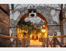 Steinbogen mit einer Glocke darüber, der in einen Innenbereich führt, der an eine historische Dorfkulisse erinnert. Der Bereich ist beleuchtet, mit Palmen, gepflastertem Boden, Holzbalken und Laternen, umgeben von Gebäuden im Fachwerkstil.