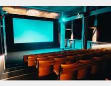 Ein moderner Kinosaal mit hölzernen Kinositzen in mehreren Reihen, einer großen Projektionsleinwand vorne und einer blau beleuchteten industriellen Decke mit Stahlträgern. Die Wände sind teilweise mit dunklen Vorhängen versehen, die eine schlichte Atmosphäre schaffen.