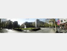 Weitläufiger Platz mit einer zentralen Grünfläche, in der ein großer Brunnen mit einer Wasserfontäne steht. Umgeben von modernen, mehrstöckigen Gebäuden mit verspiegelten Fassaden; Bäume und gepflasterte Wege rahmen die Szene ein.