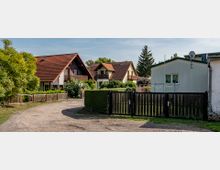 Eine ländliche Szene mit mehreren Einfamilienhäusern, die rote Schrägdächer und Balkone haben. Vor den Häusern befinden sich Holz- und Metallzäune sowie eine unbefestigte Zufahrtsstraße, umgeben von Bäumen und Hecken.