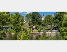 Uferbereich mit kleinen Häusern und Gärten, umgeben von dichtem Grün; im Hintergrund erhebt sich ein Kirchturm über den Baumkronen.